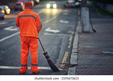 Man sweep road with broom, clean city road and roadside at night. Janitor cleanup city from garbage. Municipal worker in orange uniform sweeping street, night work