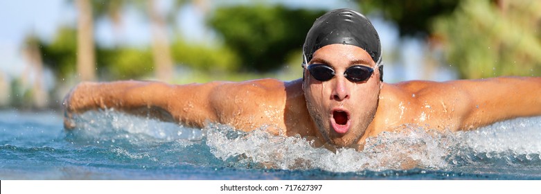 Man Swimmer Swimming Butterfly Strokes In Pool Competition Training For Triathlon. Professional Male Sport Athlete Swimmer Wearing Swimming Goggles And Cap Coming Out Of Water Portrait.