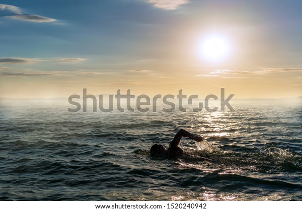 Man swimming\
in open water during a misty\
sunset