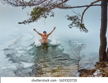 Man in swimming trunks stands in the ice hole under a pine and smiles, raising his hands