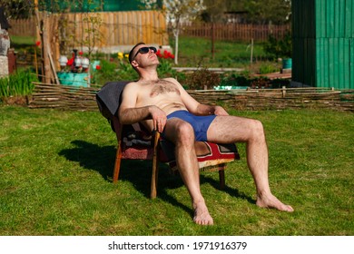A man in swimming trunks sunbathes on a ancient armchair in the yard of a country house