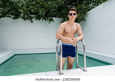 Man In Swimming Trunks And Sunglasses Standing Near Pool Ladder