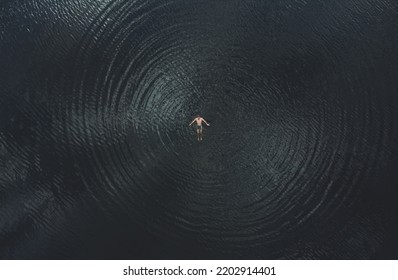 A man swims on his back in dark water. View from above. Human provokes circular waves on the water. Clouds are reflected on the surface of the water. Top view. - Powered by Shutterstock