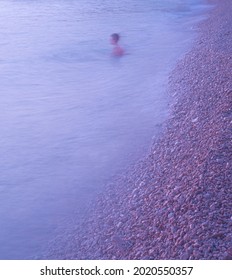 A Man Swims In The Turbulent Sea In The Early Evening At Sunset. Slow Shutter Speed Photo. Beautiful Small Sea Stones Lit By The Last Rays Of The Sun.