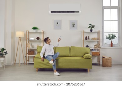 Man Switching Air Conditioning Modes Sets Comfortable Temperature While Enjoying Fresh Air At Home. Man With Remote Control Relaxing Sitting On Sofa In Living Room. Concept Of Air Conditioning System.