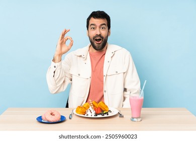 Man at a table having breakfast waffles and a milkshake surprised and showing ok sign - Powered by Shutterstock