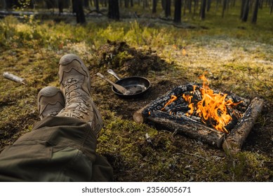 A man in tactical boots sits next to a fire in the woods. View of legs and fire. - Powered by Shutterstock