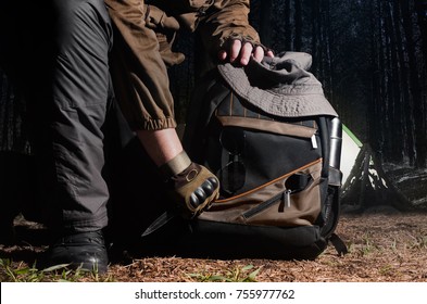 Man in tactical outfit holding a knife and kneeling for backpack with camping and tactical gear on night forest background.