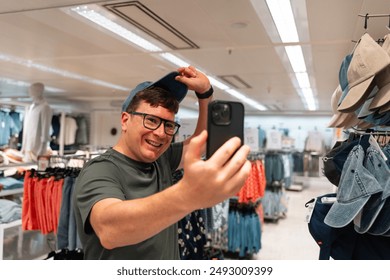 A man takes a selfie with his smartphone, trying on a new hat in a clothing store. Male consumer, second hand shopping, video call with friends  - Powered by Shutterstock