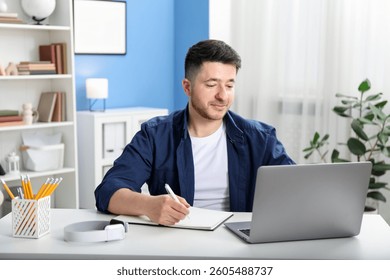 Man taking notes during online lesson at desk indoors. Self-study