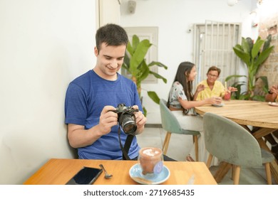 A man is taking a picture with a camera while sitting at a table with a cup of coffee. There are two other people in the background, one of whom is holding a drink. The scene is casual and relaxed