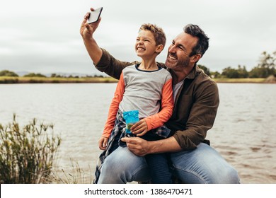 Man taking a selfie with his son sitting near a lake. Smiling father and son having fun taking a selfie on a leisure trip to a lake. - Powered by Shutterstock