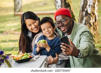 A Man Is Taking Selfies Of His Family In Nature In Front Of The Van, At The Dining Table While The Mother Holds Their Boy Who Is Eating A Healthy Snack. They Are Posing And Smiling At The Camera.