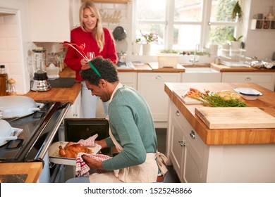 Man Taking Turkey Out Of Oven As Couple Drink Wine And Prepare Dinner On Christmas Day