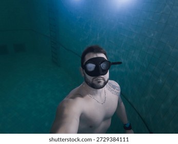 Man taking an underwater selfie while swimming in a deep pool wearing a black diving mask and a silver chain.