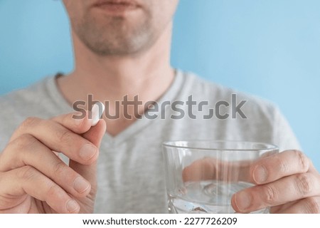 Man taking white pill of statin medicine to treat high cholesterol with glass of water on blue background. Taking medicine, health care, pharmacy and treatment concept. Selective focus