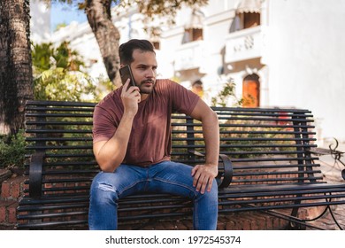 Man Talking On The Cell Phone, Showing Happiness Sitting On A Bench In The Colonial City Of Santo Domingo