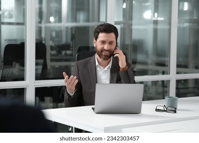 Man talking on phone while working with laptop at white desk in office