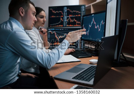 Man talks and shows on the numbers on monitor with hand. Team of stockbrokers are having a conversation in a office with multiple display screens.