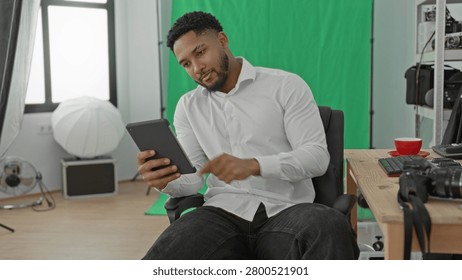 Man tapping tablet in studio beside camera gear and green screen, seated in chair with keyboard and cup on desk; concentration creativity.