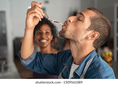 Man tasting spaghetti pasta while smiling woman look at him. Closeup face of young man tasting meal while cooking at home. Handsome guy eating noodles with fork in kitchen and feel his recipe.