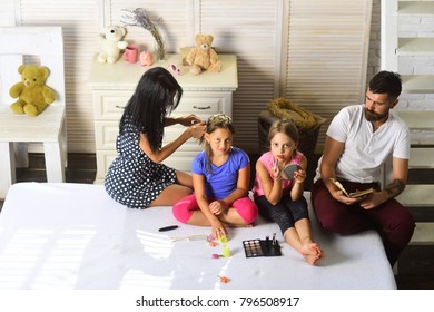 Man With Tattoo, Woman With Long Hair And Kids With Smiling Faces. Family And Fashion Concept. Family Sits In Stylish Bedroom. Dad With Beard Reads, Mom Makes Her Daughter Hairstyle, Girls Do Makeup.