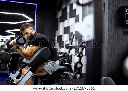 A man with tattoos performs bicep curls with a dumbbell in a sleek, modern gym. The space is well-lit and features various gym equipment against a stylish backdrop.