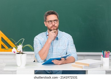 Man Teacher Thinking. Portrait Of Smart Young Tutor In Glasses With Book Think On The Blackboard In Class At School Or Collage. Male Student Study In University.