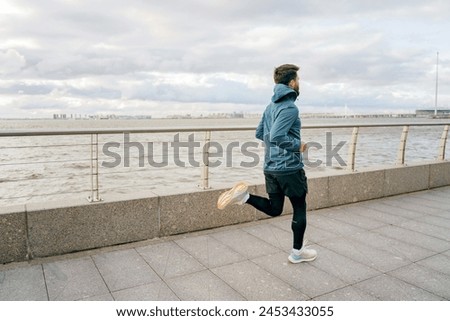 A man in a teal jacket keeps pace along the river walk, under a dramatic sky suggestive of impending weather.