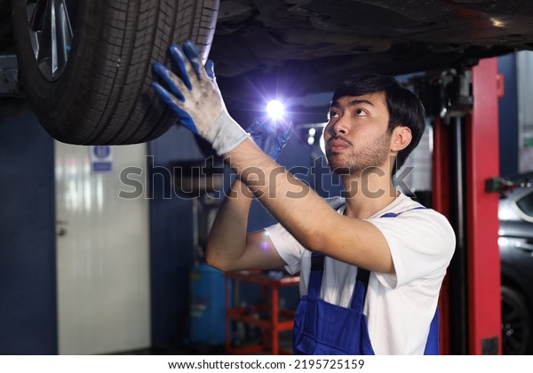 Man technician car mechanic in uniform check or\
maintenance a lifted car service at repair garage station. Worker\
holding wrench and fixing vehicle with flashlight. Car center\
repair service concept