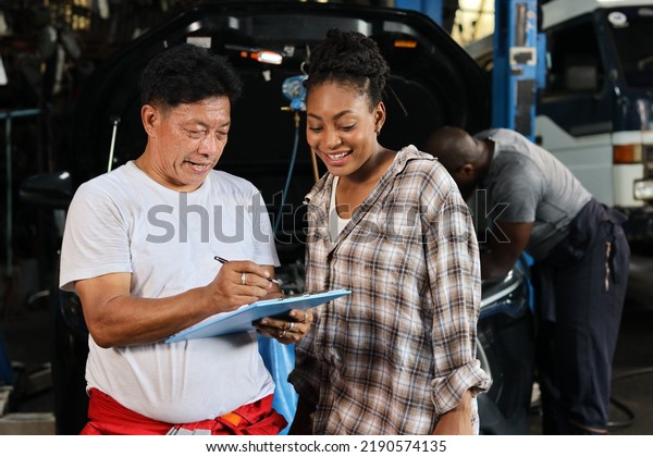 Man technician car mechanical in half\
uniform show car maintenance service report on clipboard at repair\
garage station. Auto mechanic give customer discussion on her\
vehicle repair\
problems condition