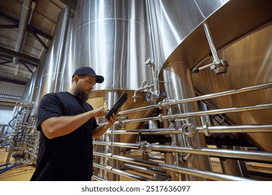 Man, technologist with tablet in hand checking the brewing system, ensuring each step of process meets strict quality standards. Concept of beer, brewery, manufacture, quality control - Powered by Shutterstock