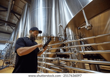 Man, technologist with tablet in hand checking the brewing system, ensuring each step of process meets strict quality standards. Concept of beer, brewery, manufacture, quality control