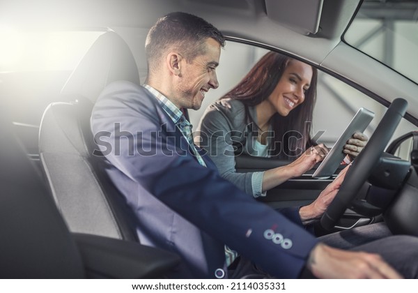 Man testing a car in the sales shop\
smiles looking into a tablet in the hands of the female car dealer\
showing technicalities.