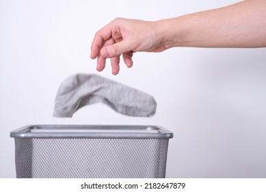 Man Throwing An Old Sock With His Hand Into A Trash Can Against A Gray Background. The Concept Of Old Clothes.