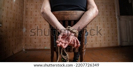 A man tied with a rope sits on a chair in an abandoned room