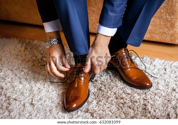 A man ties up his shoelaces on\
his brown shoes in the room. Blue suit and patent leather shoes.\
