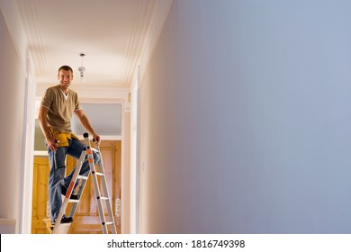 Man With Toolbelt Doing DIY At Home Standing On Step Ladder Below Ceiling Light Fixture And Looking At Camera Happily.