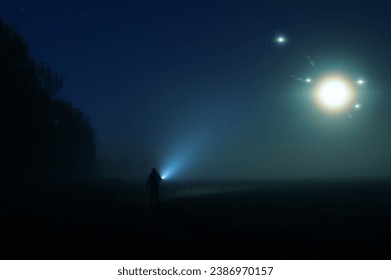A man with a torch standing in the countryside on a misty summer night. With glowing UFO lights floating in the sky.