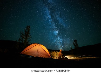 Man Tourist Near His Camp Tent At Night Under A Sky Full Of Stars. Orange Illuminated Tent.