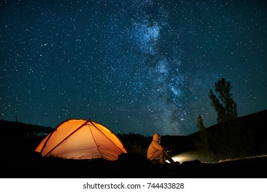 Man Tourist Near His Camp Tent At Night Under A Sky Full Of Stars. Orange Illuminated Tent.