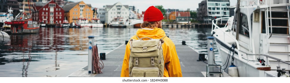 Man tourist with touristic rucksack wearing yellow jacket walking among authentic fishing boats. Hipster traveler wanderlust with backpack. Wide image
