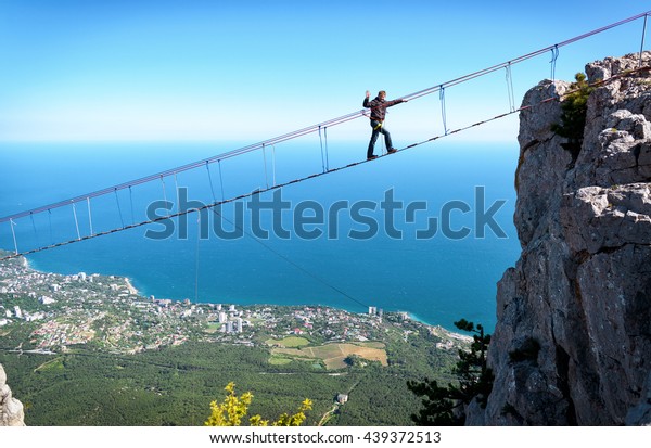 Man tourist walks on rope bridge at Mount Ai-Petri,\
Crimea, Russia. Amazing view of high rope bridge over Black Sea.\
Mountain, climb, skyline, hike, travel and tourism theme. Crimea -\
May 19, 2016