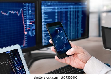 Man trader sitting at desk at office in front of monitors with stocks data holding smartphone browsing application monitoring price changes on candle bar online using digitla devices close-up