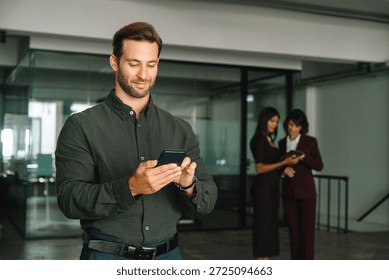 Man trader using digital technology cellphone phone mobile for business trade work. Focused young Latin businessman holding smartphone telephone device standing in office with people on background