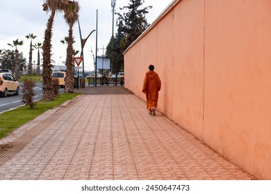 man in traditional long attire walks down street along high wall Red, City Marrakech, back view, authentic urban African landscape, daily activities people - Powered by Shutterstock