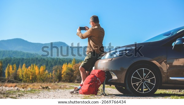 Man travel by car and making photo and video\
on mobile phone. Break between road trip. Mobile phone apps for car\
owners. Man using rent car through mobile apps. Beautiful nature\
with mountaines.