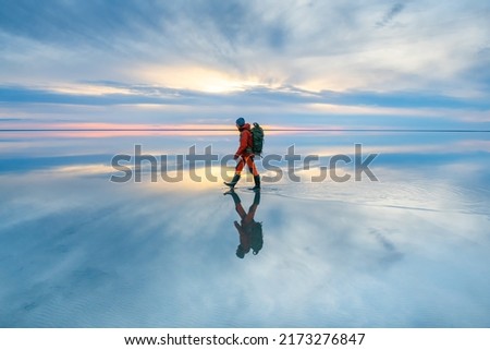 Man traveler with backpack walking on the salt lake at sunset. Sky with clouds are reflected in the mirror water surface. Travel and adventure concept
