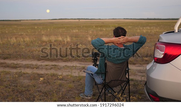 A man traveler in car stopped at a camping in\
evening and takes pictures with a camera next to car. Freelance\
photographer working outdoors, beautiful moments for photos and\
videos. Driver is resting