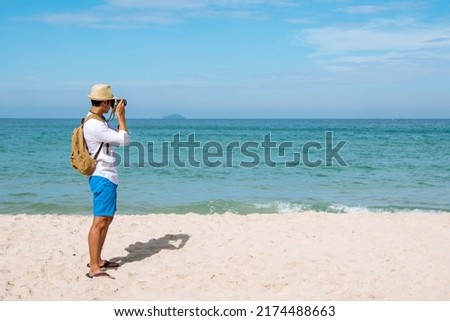 Man traveler using digital camera on summer beach blue sky take a photo. Asian Photographer man journey relax on the beach in summertime. Man backpack shooting photo asia destination holiday trip
